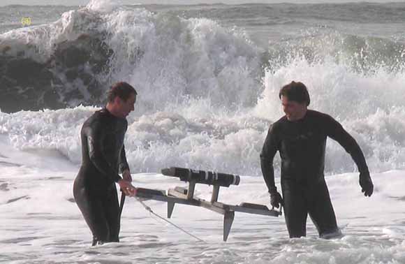 photo of two scientists in wetsuits carrying equipment through the surf