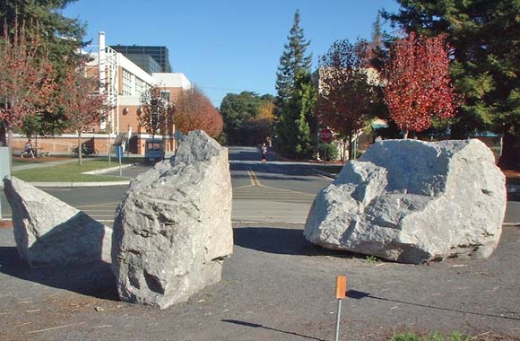 Photograph of three large, light-colored boulders surrounded by gravel. A circular brass benchmark is visible in the concrete sidewalk in front of the boulders.