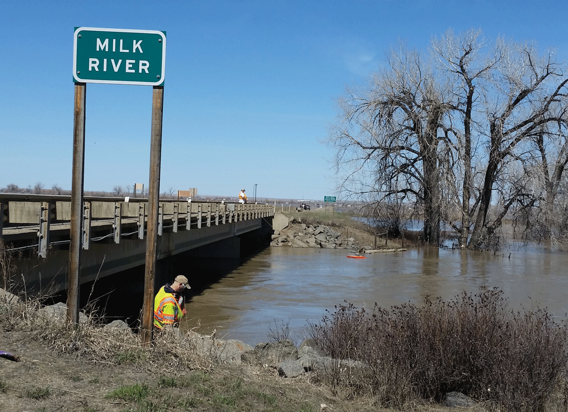 Photograph of Milk River taken from a bank near a bridge. Scientists are sampling
                     the river.