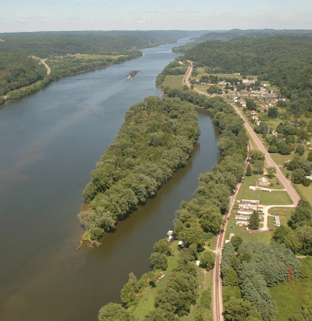 Photograph shows view of river with a small, wooded island near the bank. On the bank,
                     buildings and roads can be seen.