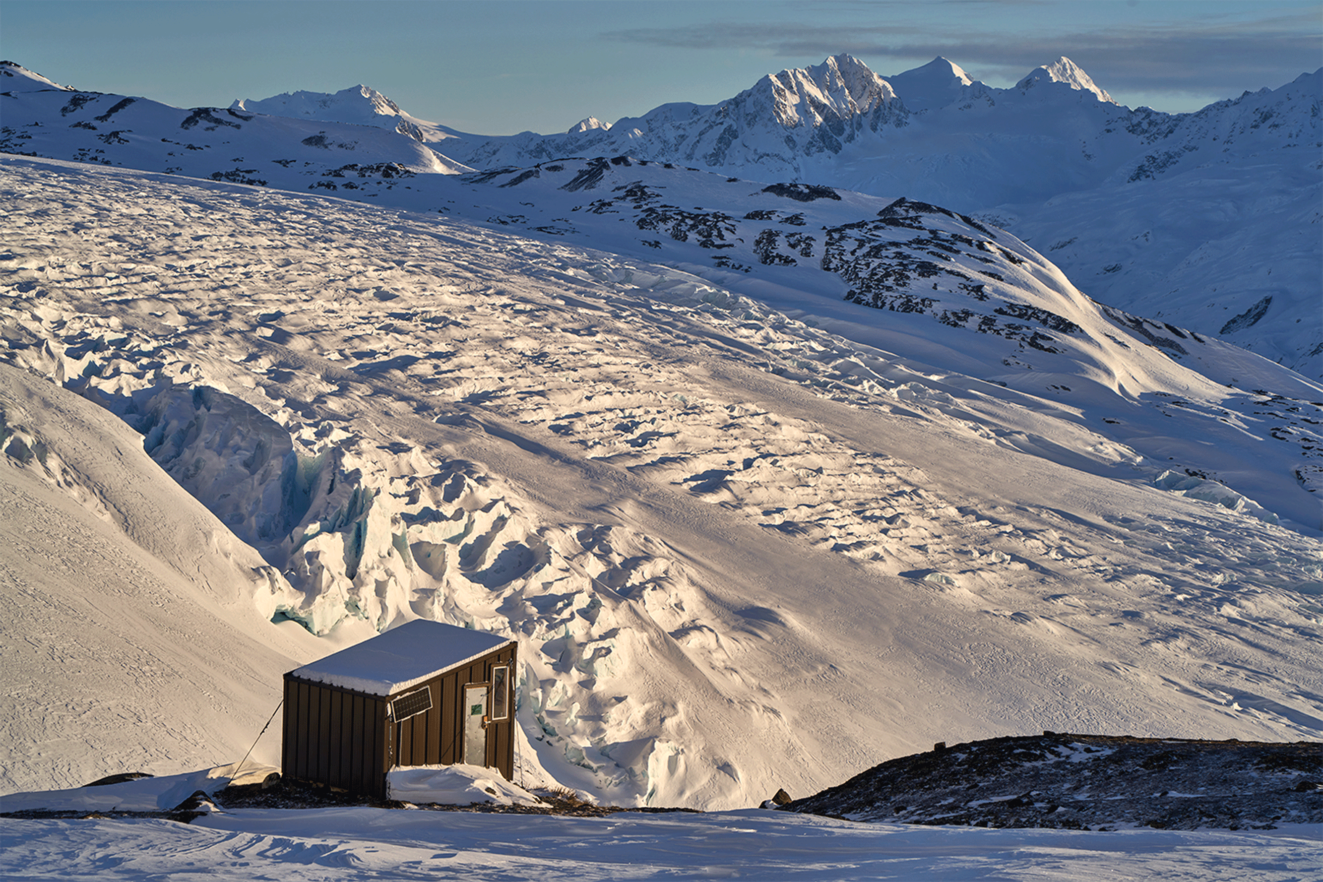 A small hut overlooks the Wolverine Glacier and distant snow-covered peaks in Alaska.