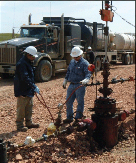 Workers collecting sample at an oil well pad, with a truck shown in the background.