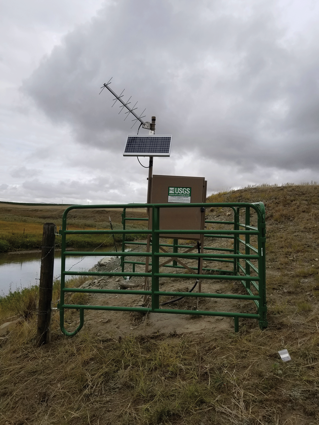 Photograph of streamgages. A, streamgage surrounded by green metal fence next to river.
                     B, same from different angle.