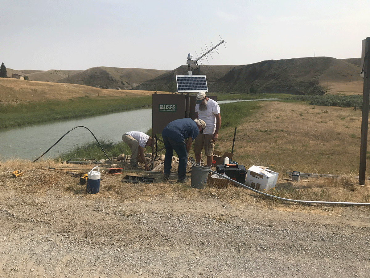 Photograph of streamgage next to river, with three scientists standing next to the
                     streamgage