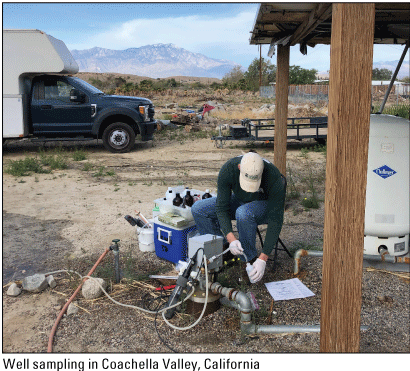 A USGS scientist collecting a sample from a well in Coachella Valley, California.
                     There is a mountain in the background, and the sky is partially cloudy.