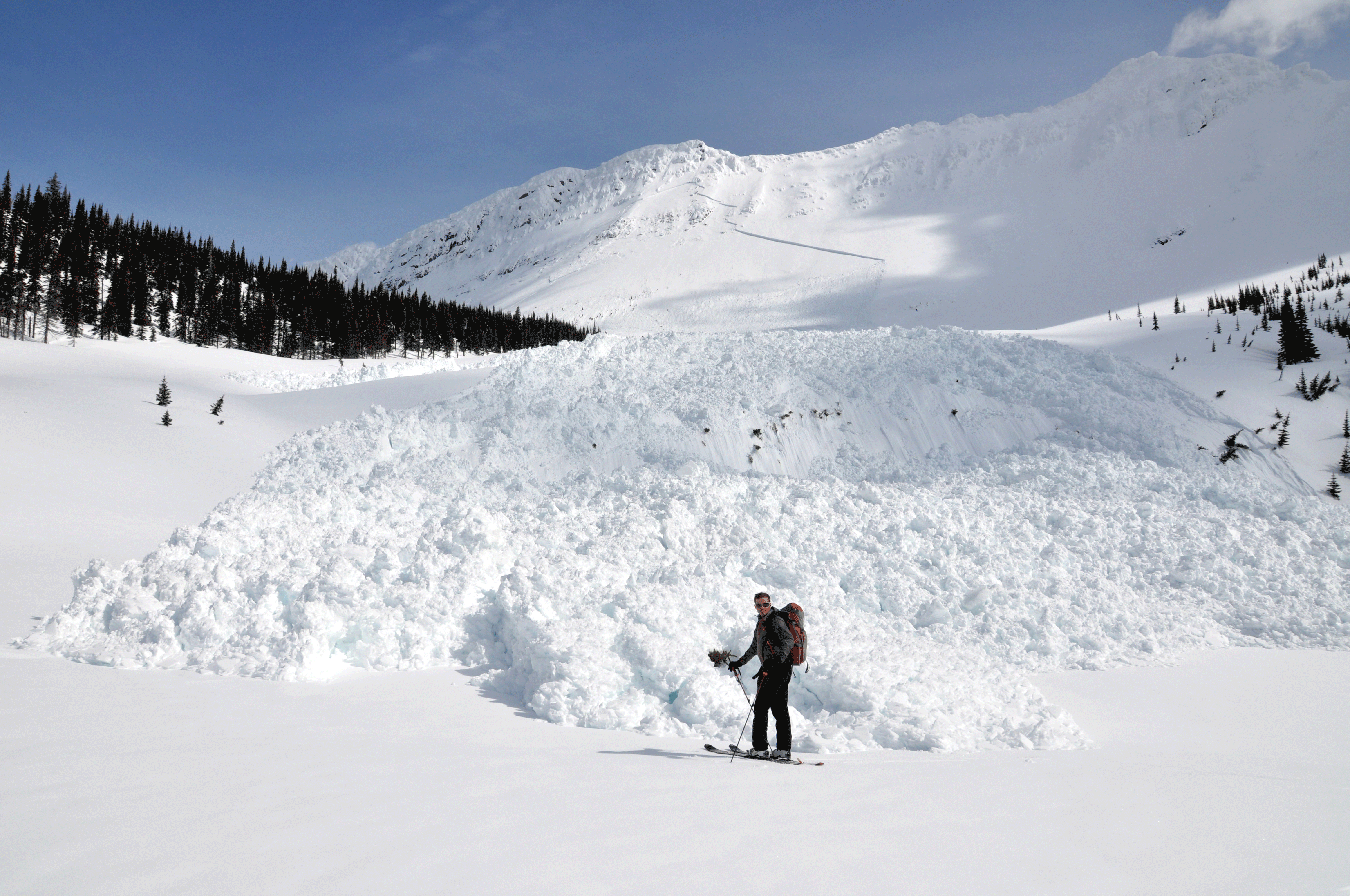 Person in ski gear standing in the snow in front of a large avalanche debris deposit.