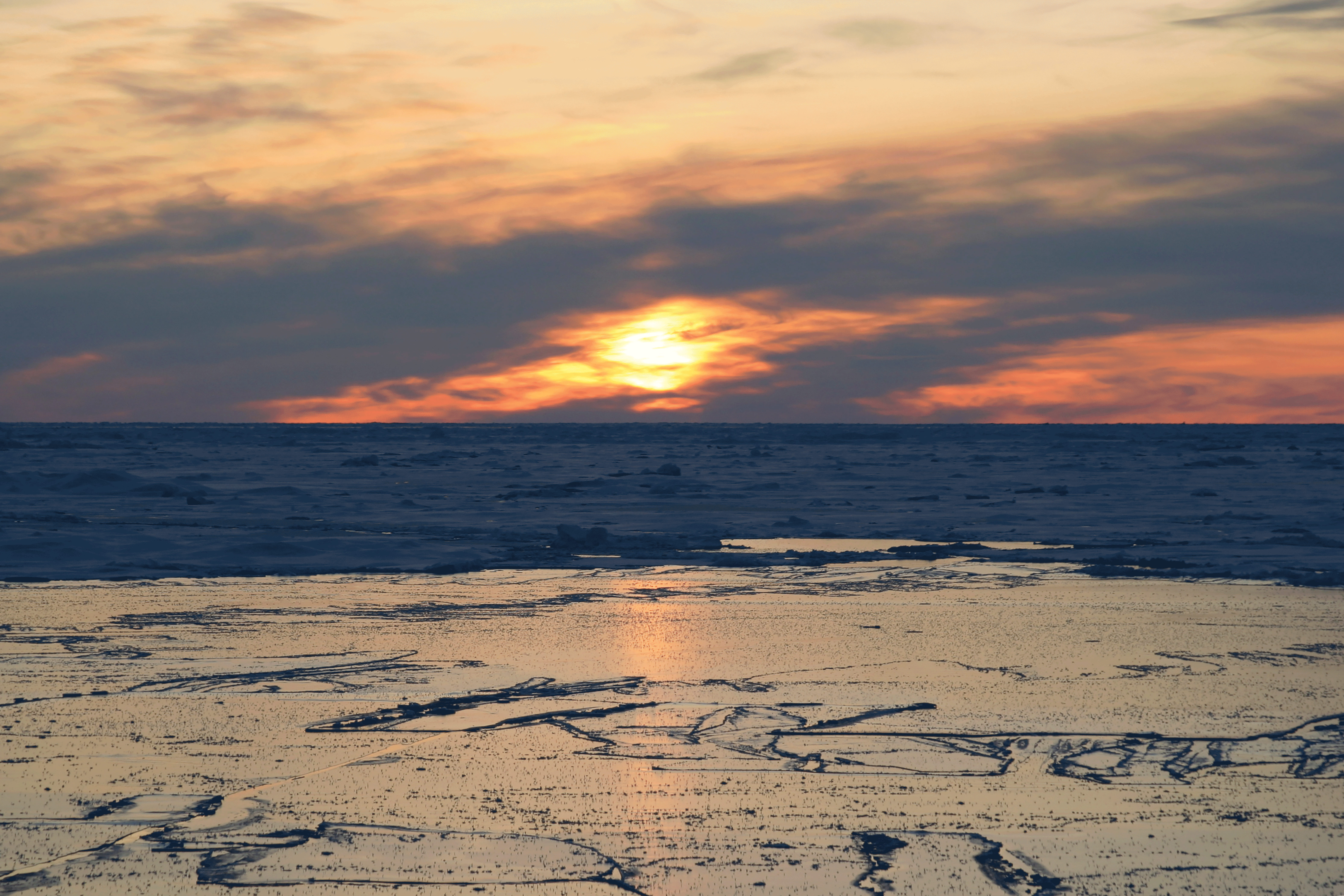 Cracked sea ice floats in the foreground in front of an overcast sunset.