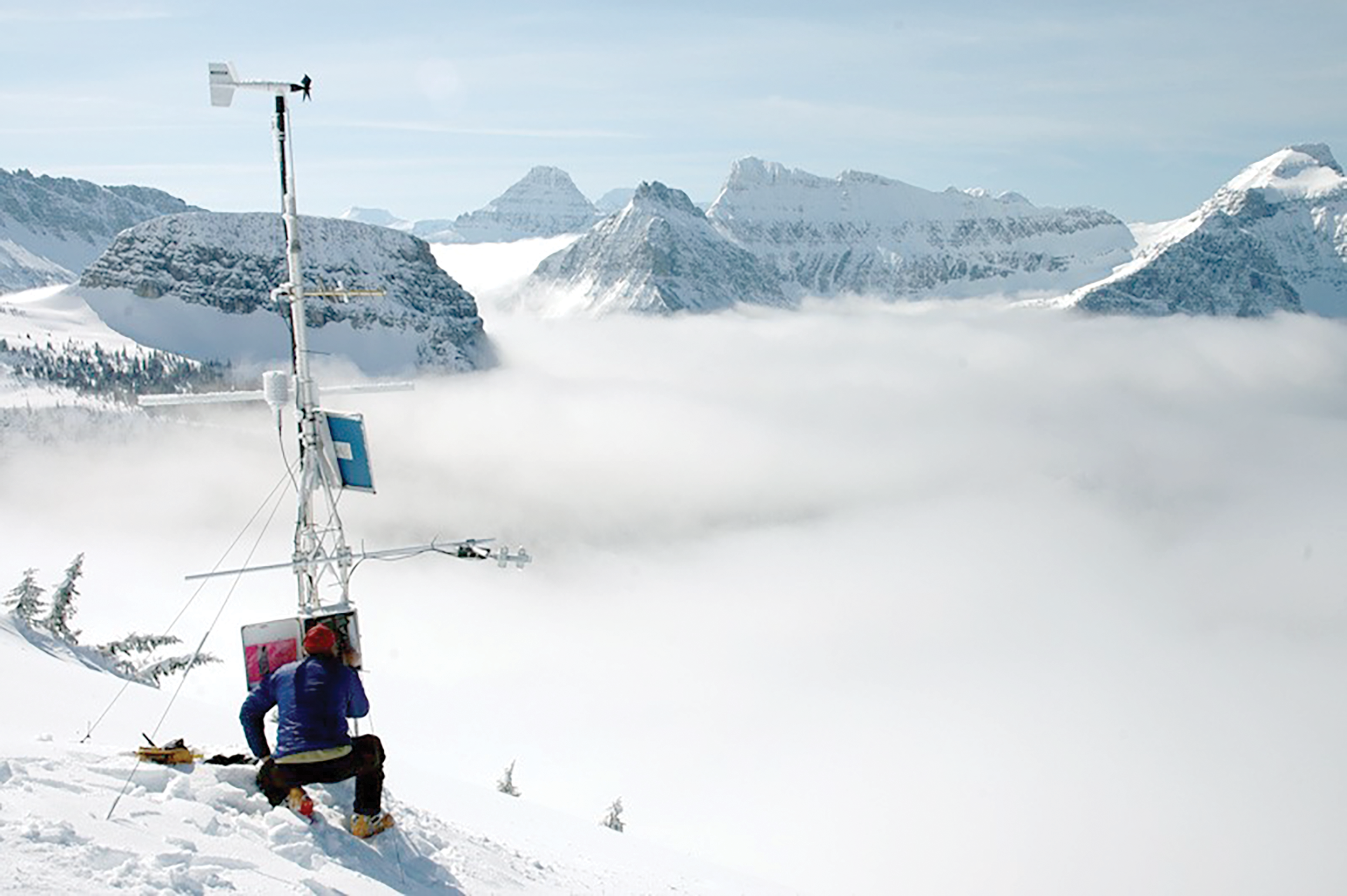 Person kneeling in snow in front of the weather station surrounded by mist and mountains
                        in the background.