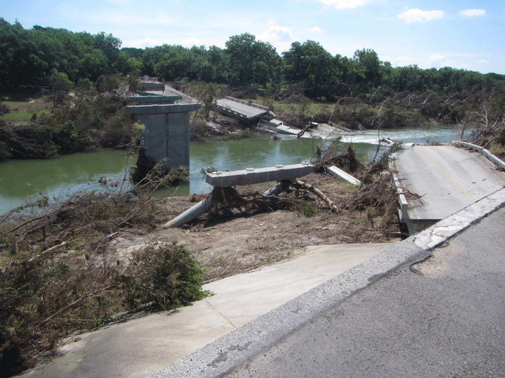 Image depicts destroyed bridge, with large pieces of debris in the river and on the
                     surrounding riverbank. On the left side of the riverbank, there is a green landing
                     lined by trees.  