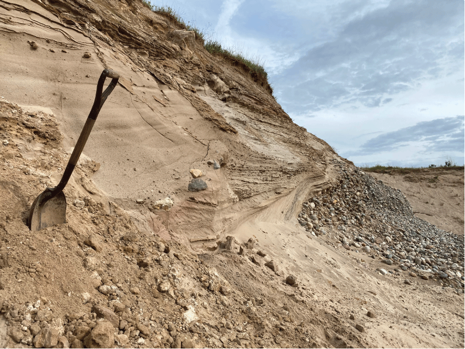 A sloping, exposed deposit with a shovel stuck into it in the foreground. A cloudy
                        daytime sky is shown.