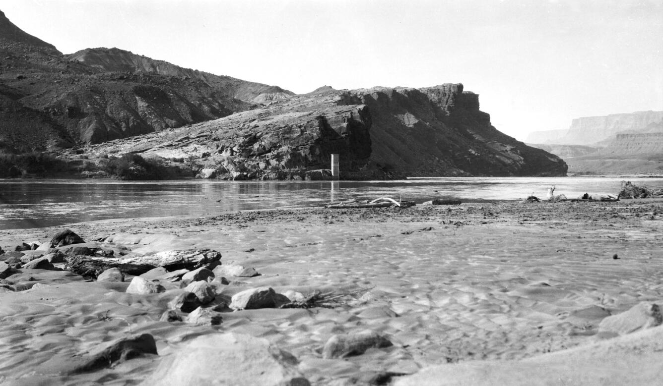 A black and white photograph shows the Lees Ferry streamgage located under cliffs
                     on the far side of the Colorado River, with a sandbar on the river's near shore in
                     the foreground.