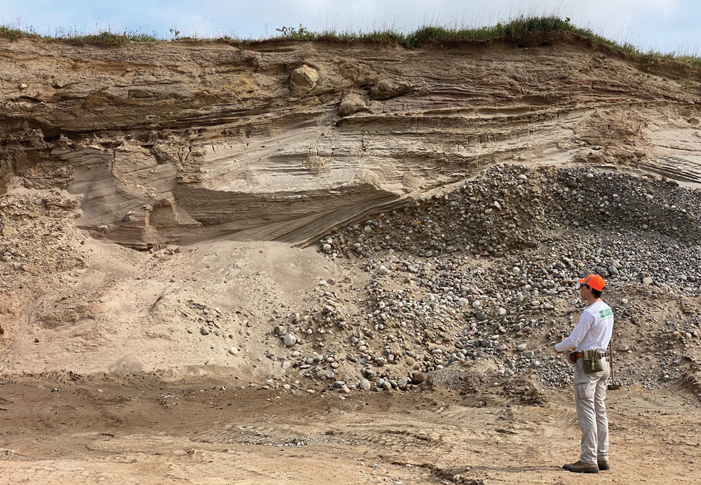 A scientist stands in front of and to the right of a large outcrop of glacial sediment
that contains layered sand beds above a layer of gravel.