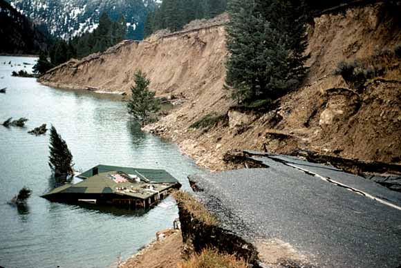 Photograph of house nearly submerged with broken-off road in foreground and landslide cliff in background