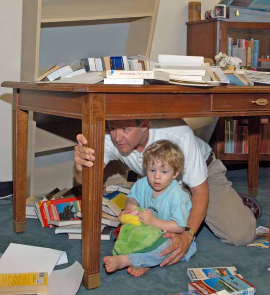photo of father and little boy holding onto a table leg.  A bookcase has tipped against the table and there are books scattered all over the floor.