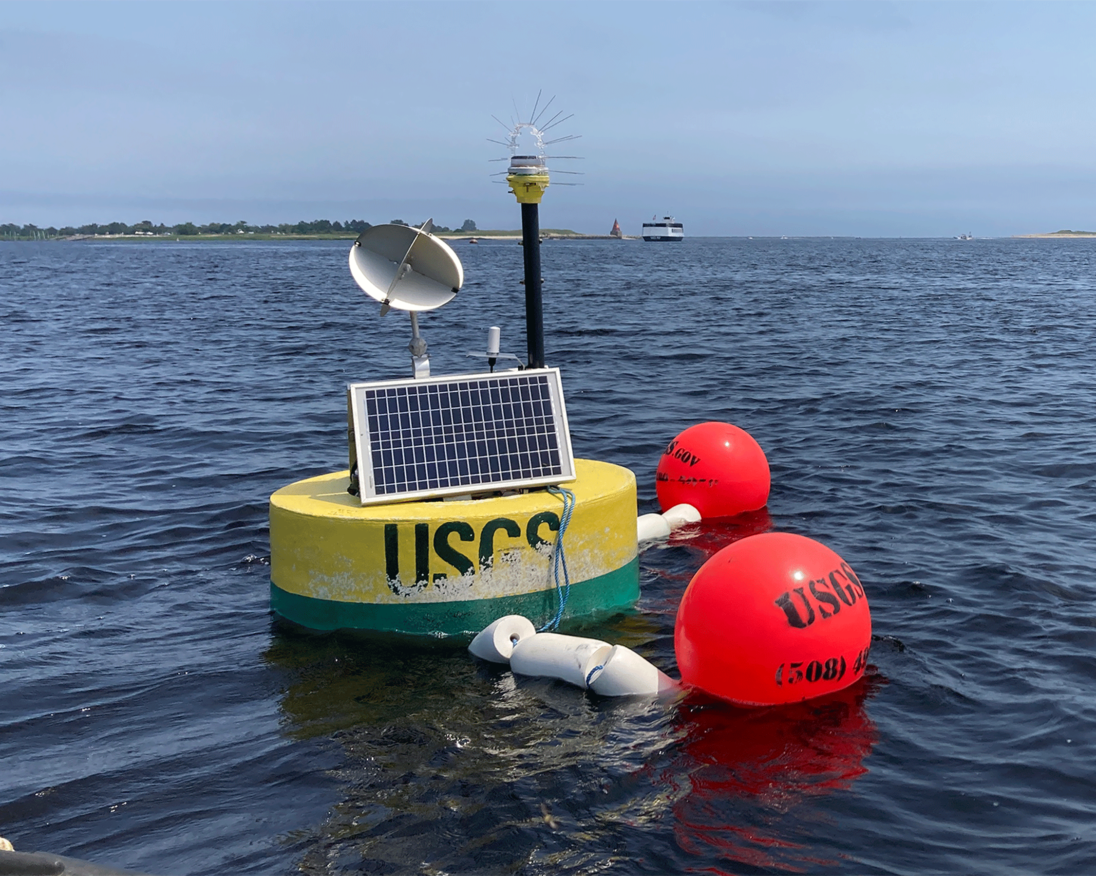 Photograph of a small yellow buoy marked “USGS” floating in a wide bay, outfitted
                     with a solar panel, lights, equipment, and high-visibility floats.