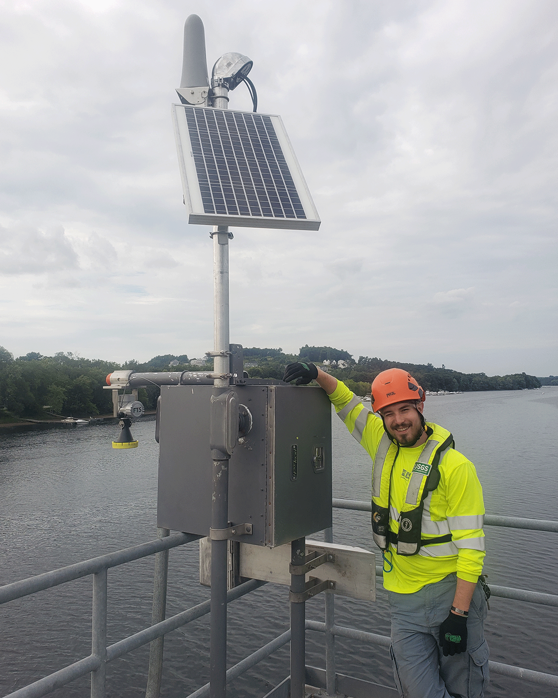Photograph of an employee wearing high-visibility clothes and a helmet. He is above
                     a water body, smiling, and leaning on a metal box with a solar panel.
