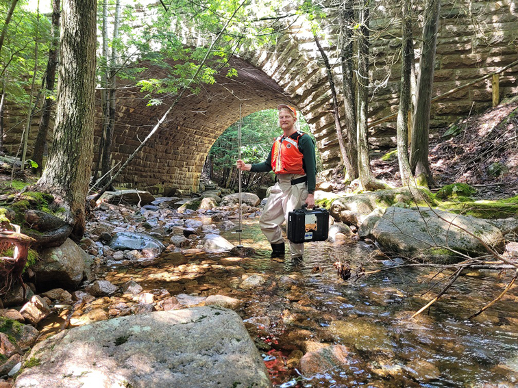 A man in a life vest and waders, smiling while holding measurement equipment, stands
                     and poses in a small, wooded stream. In the background is a large, arched stone bridge.