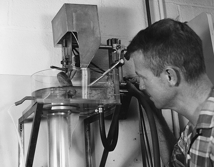 Dr. John S. Schlee, USGS, prepares to drop a sample of sand (adhered to the upended disc at center) into a long plexiglass tube in order to make a size analysis of the sand.  The weight of the particles and the time it takes them to fall down the tube is measured by a transducer and recorded as a "size curve" on tape.  This work is part of a joint WHOI-USGS study on the Atlantic Continental Shelf and Slope.