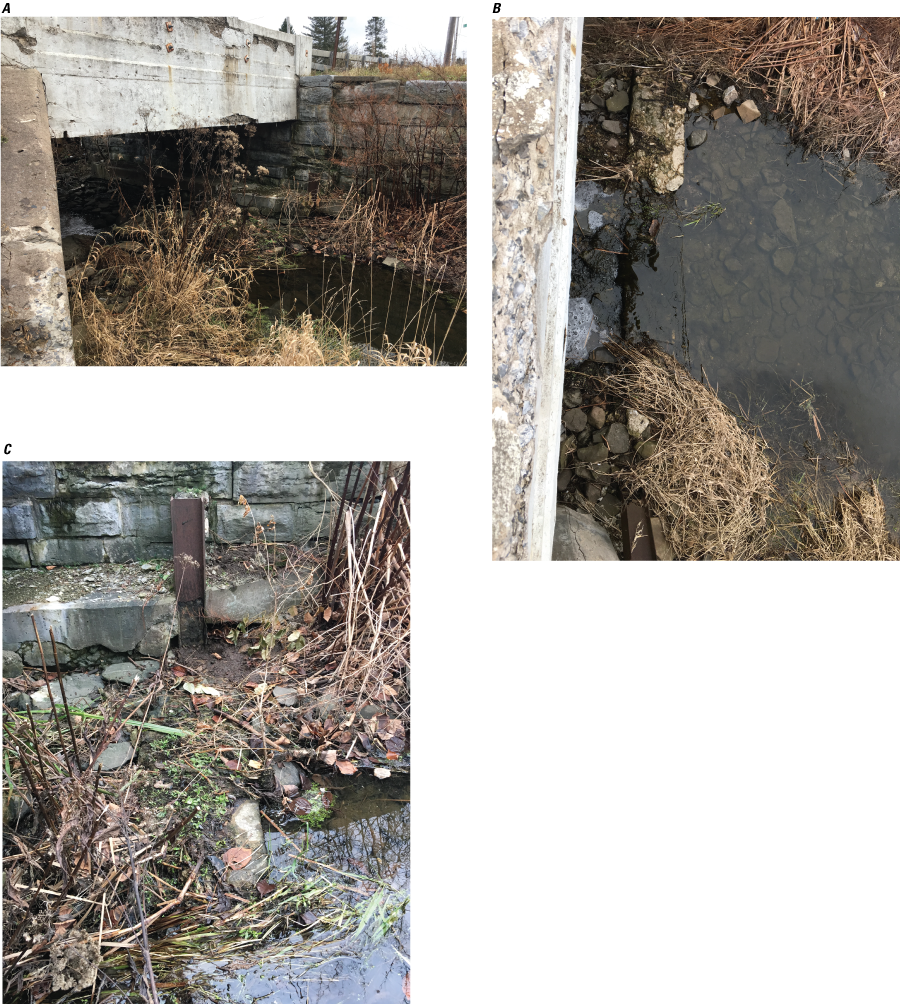 A small stream with a few inches of flow passes underneath a white-painted concrete
               bridge. In the ditch, there is broken concrete and I-beams.