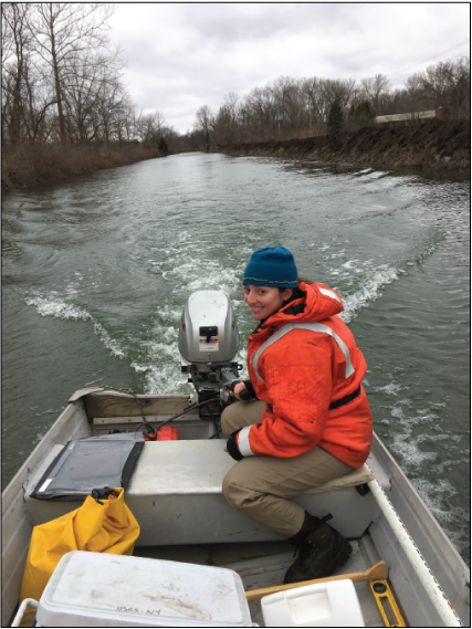 A smiling technician in a bright orange coat controls an outboard motor on an aluminum
                        boat, leaving a wake behind them in the canal.