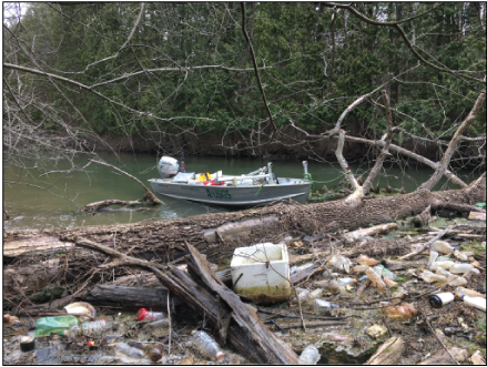 A large, dead tree is resting across the canal. An aluminum boat with USGS on the
                        side is tied to the tree. On the upstream side, there is a floating mat of hundreds
                        of plastic bottles.