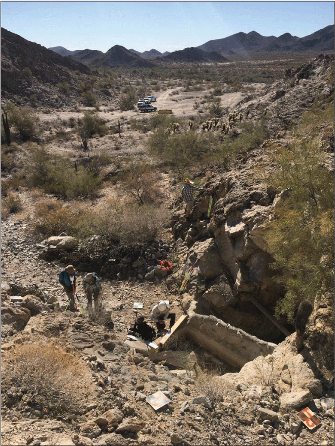 2.20. Surveyors work within a cobble-bed stream near a rectangular catchment basin
                  beside the tank entrance. In the distance is a sandy arroyo valley.