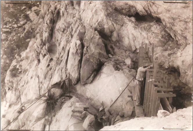 2.29. A scaffolding-like frame spans the mouth of a rocky channel. A person is on
                  the frame and two other people are working on a rock ledge below.