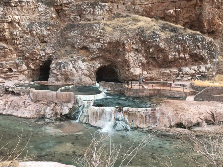 A rock face with two cavern entrances is behind several pools that cascade water into
                  the adjacent river set slightly below.