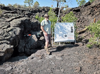 Researcher standing next to a poster with a frame on a rocky slope with light foliage
                  near the Bonito Flow Ice Cave entrance.