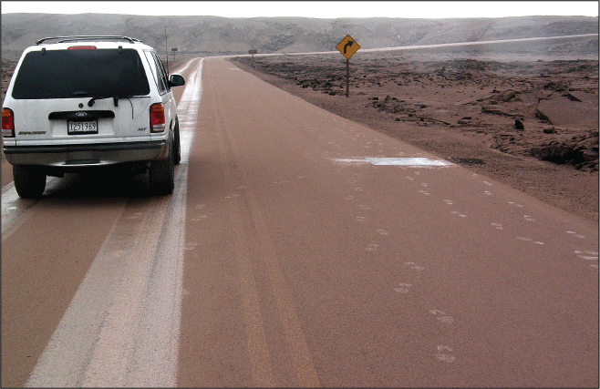 Alttext 15: There is a car parked on a road. The road is covered in a thin layer of
pinkish-brown ash, in which there are footprints and tire tracks.