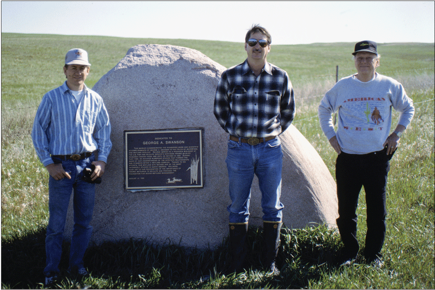 Three men standing in front of dedication plaque hung on a large rock with grassy
                     area in background.