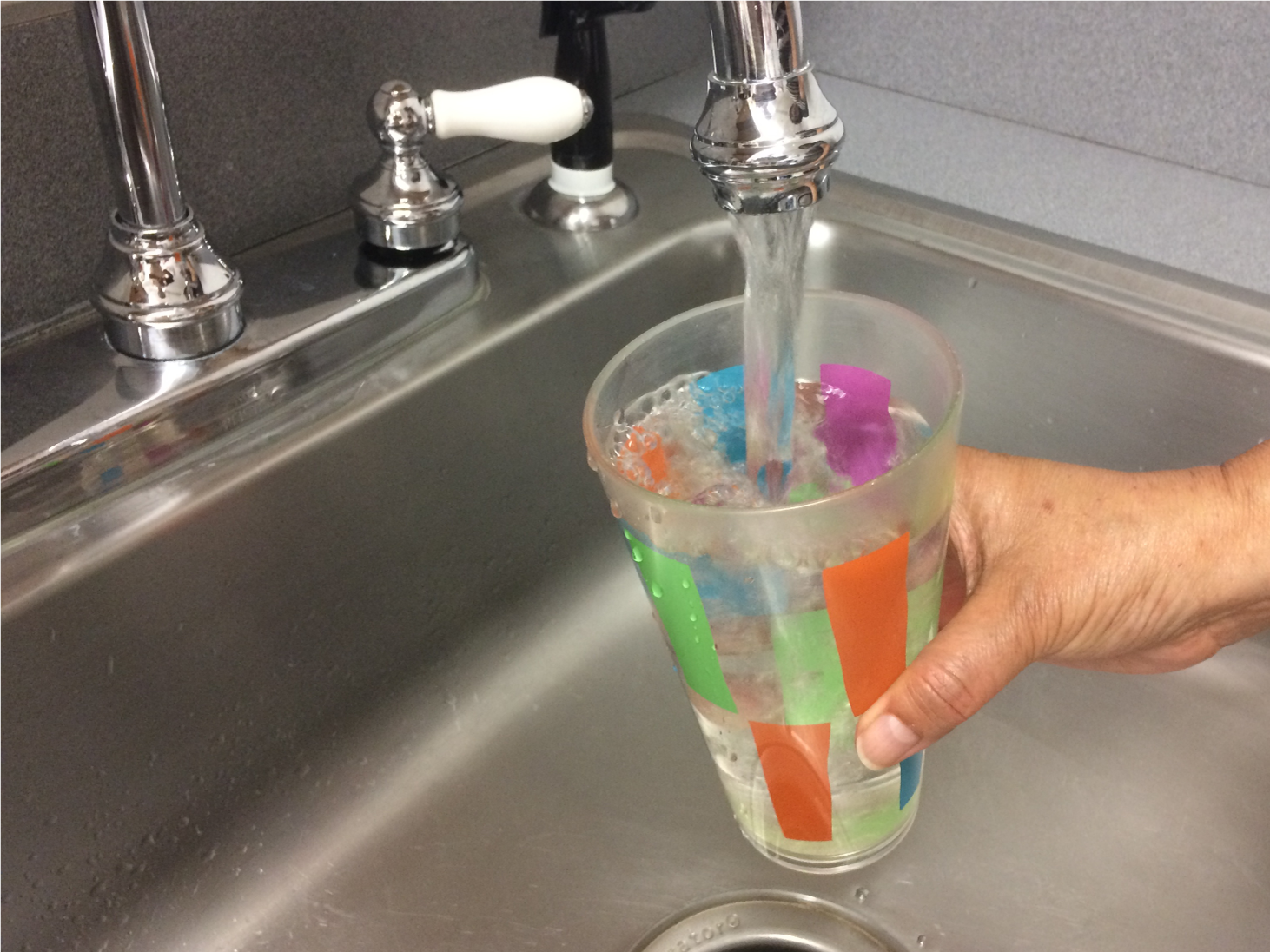 Photograph showing a hand holding a drinking water glass under a kitchen sink tap
                        and tap water filling the glass.
