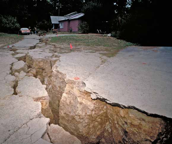 photo of deep, wide crack in the Earth with an intact house, trapped car, and wrecked driveway in the background