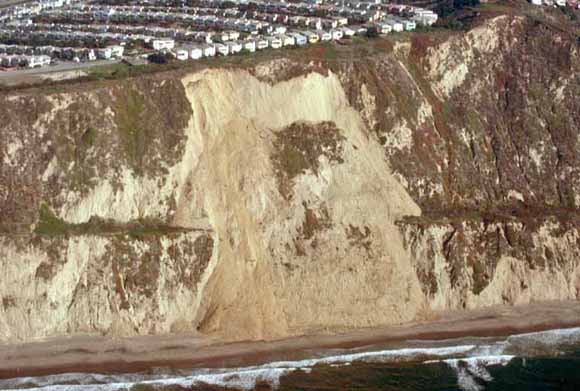 photo of landslide along coastal bluffs.  A large housing tract is situated right on top of the bluff.  The waves are crashing at the bottom of the bluff
