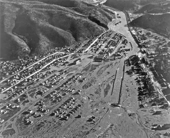 old black-and-white photo taken from a low-flying airplane showing flooded town along riverbank