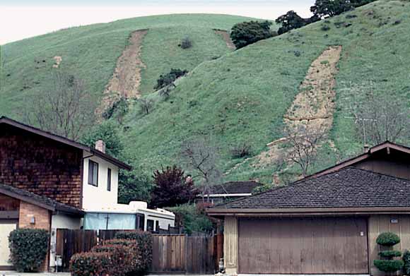 photo of houses in the foreground with a hillside in the background; the hillside has two landslide scarps in it.