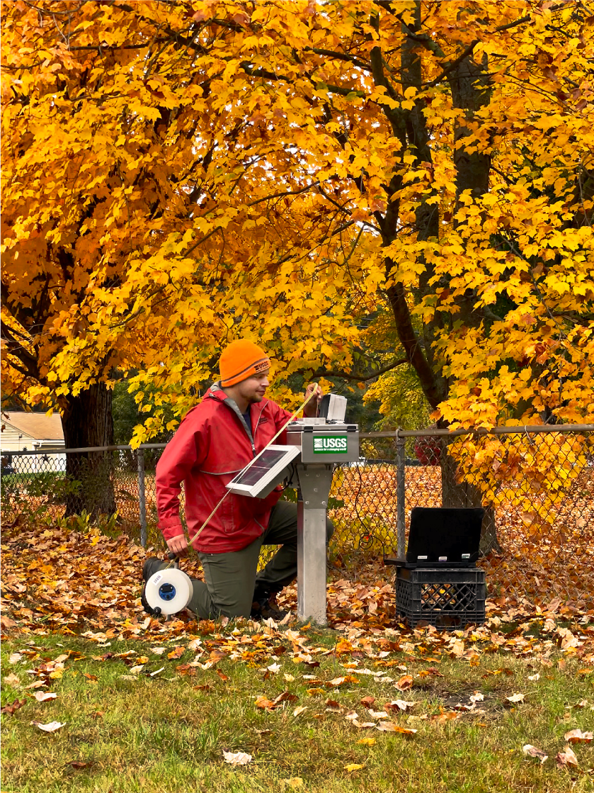 Scientist with tape measure and instrument measuring groundwater level; with fall
                        leaf foliage in the background.