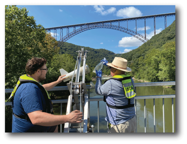Two USGS personnel removing bagged water from a DH-2 sampler attached to a bridge
                           crane.