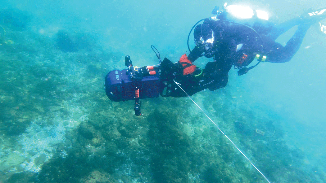 A self-contained underwater breathing apparatus (SCUBA) diver collecting data using
a blue underwater camera over a rocky lakebed in clear water.